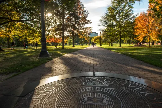 Scenic view of Ohio State campus with autumn trees, paved walkway, and university seal in foreground.